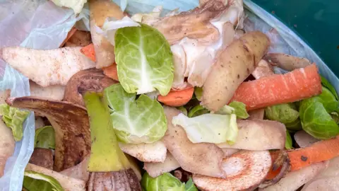 Colourful vegetable peelings in a food waste bag with brussels sprouts, potato skins, carrots and the core of a white cabbage. The bag is white and made of plastic.