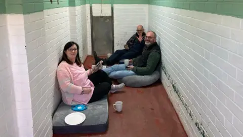 Three people - a woman and two men - sit in a small brick kennel enclosure at an animal shelter. They have dog beds and toys around them.