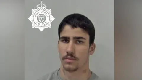 A man with dark hair and short facial hair, wearing a grey jumper, stands in front of a plain wall in a police custody photo, with the West Mercia Police crest to the top left of the picture.