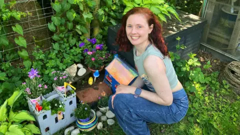 Christie Barnes, a woman with red hair wearing blue jeans, squats down next to a miniature castle and house for mice in her back garden. 