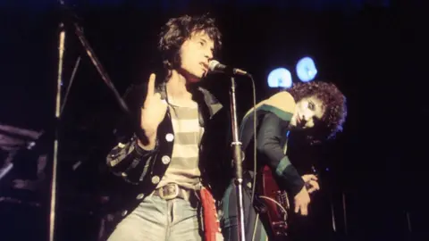Getty Images The Sensational Alex Harvey Band. Alex Harvey has dark, shoulder-length hair and is wearing a black jacket, a striped T-shirt and jeans. He is singing into a mic which is on a stand. A guitarist with dark, curly hair and face make-up is leaning over, looking at him