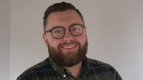 Sanderson Recruitment A smiling man with dark-framed glasses looking down the barrel of the camera. He has a thick beard and wears a tartan shirt with the top button opened. He stands in front of a white wall. 