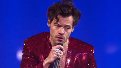 Getty Images Harry Styles on stage singing into a microphone Styles is holding while wearing a sparkling, deep red sequinned jacket over a bare chest. The background is lit in vivid blue tones, creating a dramatic concert atmosphere.