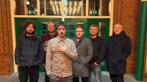 Bridge 55 (LtoR) Richard Campbell, Richard Willis, Joe Karchud, Sam Collins, Luke Bryant and Martin Davies. A group of 6 men stood in a row in a court yard, Behind them is a set of double green doors with window pannels