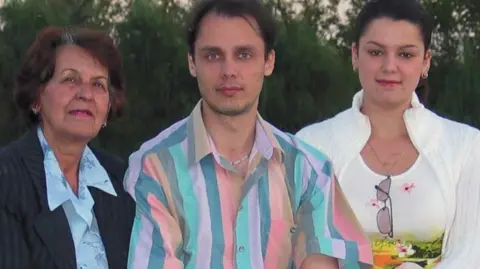 Family Three members of a family smile at the camera, with the woman on the left dressed in a dark grey jacket and a light blue blouse, the man in the middle who has dark hair is wearing a striped shirt, while the woman on the right is wearing a white wool jacket and white top.