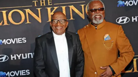 Getty Images Composer Lebo M, wearing a dark suit and white shirt, and actor John Kani, wearing a tan suit on the red carpet for Lion King.