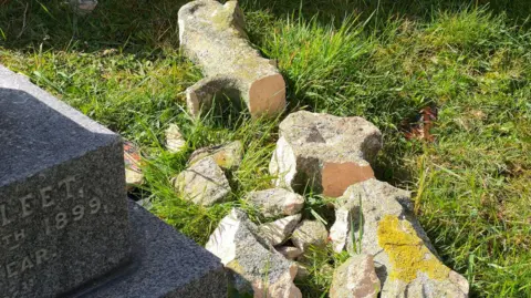 The remains of a stone cross which has fallen to the ground. The stone is covered in yellow moss. It is next to a grey memorial with an engraving denoting it dates from 1899.