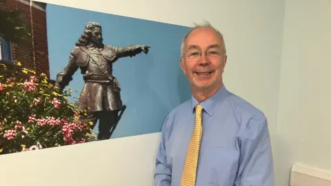 Amy Holmes/BBC A picture of Buckinghamshire Council leader Martin Tett. He is stood next to a picture of historical figure John Hampden. He is wearing a yellow tie with black dots on it and is smiling.