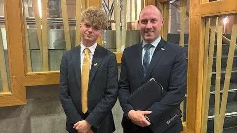 Ryan Quinn Ryan, with his distinctive curly hair, poses for photo with the health minister. He is in a dark suit and bright yellow tie. Gray wears a dark suit and dark grey tie.