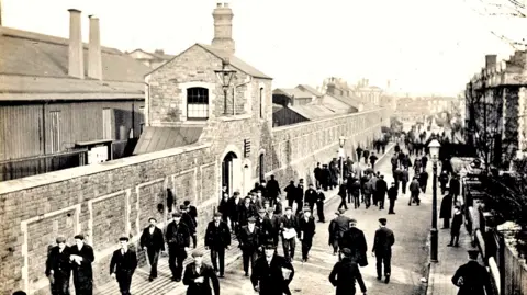 British Rail A black and white photo of a street lined by houses on the right and a long stone wall with an archway on the left. Behind the wall are large buildings, some with tall chimneys. Many men in dark coats and flatcaps are walking in the street.