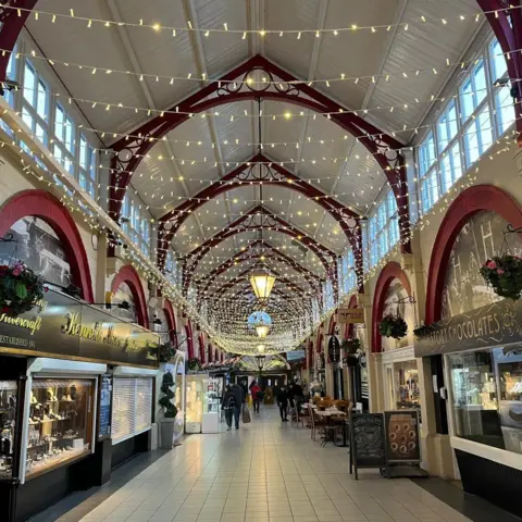 The inside of the market. There are strings of lights above the hall, which is lined with shops.