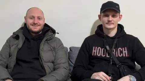 Saul and Joel are sitting on a sofa in a room, with a white wall behind them. 