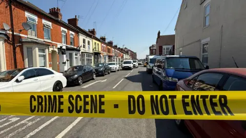 Steve Hubbard/BBC A late Victorian-style terraced street of houses with cars and buildings on either side. In front is bright yellow police tape saying "crime scene do not enter" in black block capitals. 