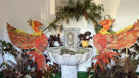 Kirsty Johnson Two large orange red and yellow phoenixes are on a wall around a stone well that is white. There are brown and green plants surrounding them and branches above them