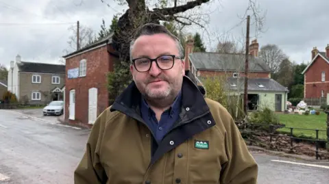 A man wearing glasses in a brown coat and blue shirt. Homes and a road are behind him.