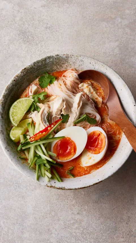Top down view of a stone coloured bowl with chicken laksa in topped with boiled eggs, veg and lime slices