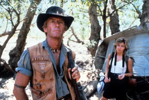 Getty Images Paul Hogan in the foreground wearing a hat, denim shirt and tan-coloured waistcoat. Linda Kozlowski is in the background, wearing a white vest and black shorts.