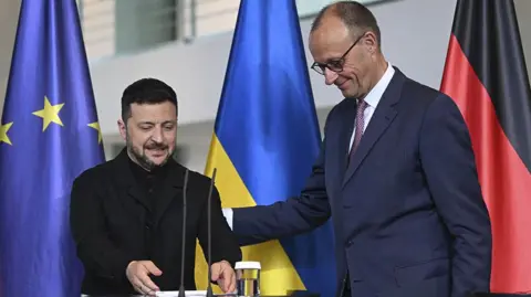 Germany's chancellor in a light blue suit on the right puts his arm on Zelensky who is wearing black