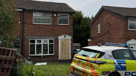 BBC The exterior of a house with a boarded up door. There is a police car parked outside. 
