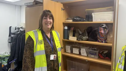 Christine is wearing a fluorescent tabard and a brownish top underneath. She has brown medium length straight hair. She is standing in a room next to a cupboard containing lost property items.