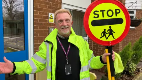 BBC A man in a green hi vis jacket holding a yellow and red lollipop stick with an image of children crossing it. 