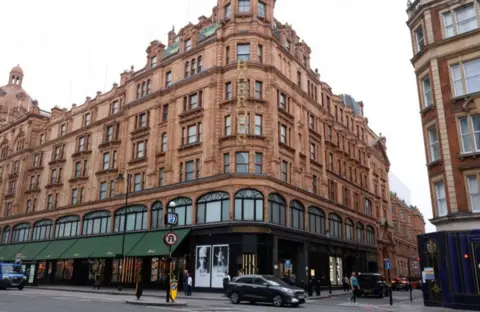 An image showing the red brick Harrods building with green awnings and a gold illuminated vertical sign, in September 2024