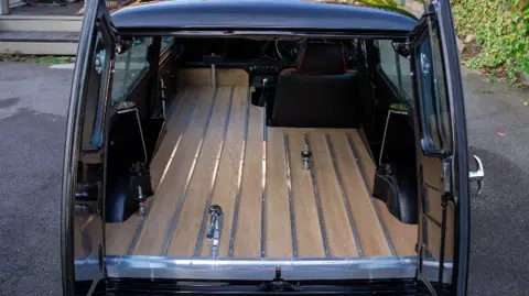 Their Last Mini Adventure A black converted Mini Clubman hearse, with rear doors open, viewed directly from behind. There is red detailing around the black driver's seat and metal features for securing a coffin.