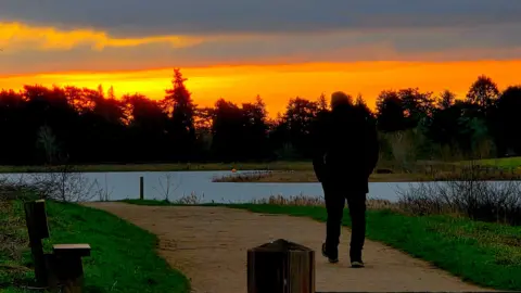 ALFIE COE/WEATHERWATCHERS A man enjoys a New Year's Day walk in Sywell, Northamptonshire, against the backdrop of the orange and yellow sunrise. He is walking on a path alongside a waterway. His back is to the camera. 