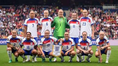 Getty Images The Lionesses - the England women's football team - pose for a photo on a pitch with a large crowd in the stands behind them.