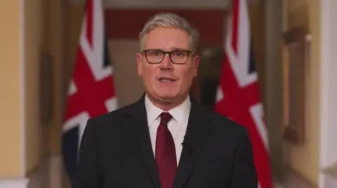 Downing Street Keir Starmer looks to camera and stands in front of UK flags as he speaks 
