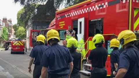 Group of firefighters with yellow helmets with a fire appliance to the right and buildings in the distance.
