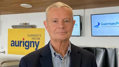 BBC A man, with short white hair, looking directly at the camera. He is wearing a blue and white stripy top and blue blazer. Behind him is an airport check in desk. 