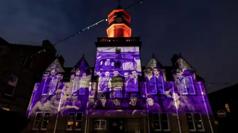 Paul Campbell The projection on a building is purple and shows a group of workmen in an archive image. There is a cab of a vintage lorry behind them.