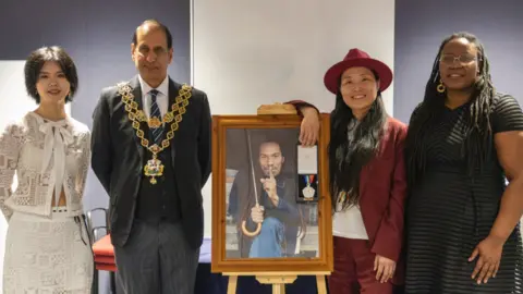 A group of people stand in room, alongside a portrait of a man who is holding an umbrella.