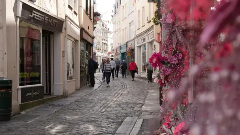 People walk along a street with shops on it in St Peter Port, Guernsey. The shop front closest to the camera has flowers outside. The street is cobbled.