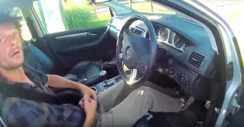 A man is captured on a body-worn camera sitting in the driver's seat of a car. His feet are in the footwell and he has his hands in his lap with his head turned to talk to the police officer. He is wearing a checked shirt and light trousers and has a blank expression on his face.