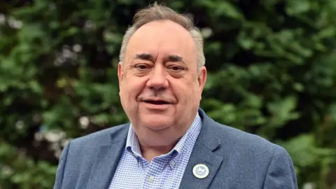 Getty Images Alex Salmond smiling in medium close up. He is wearing a blue tweed jacket, open-necked button down white and blue checked shirt and an Alba party badge