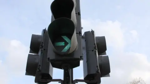 BBC A close up of traffic lights. The signal facing the camera is lit green with an arrow pointing to the right. There is a tree in a background and there are clouds in the sky.