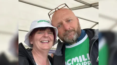 Trish Prosser Trish has short red hair and is wearing a white bucket hat. Mark is bald with brown and grey facial hair, glasses on his head and is wearing a green top which says MacMillan on it in white letters. He has a dark jacket over the top.