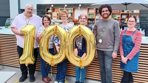 Wiltshire Council Six adults standing in a row in an indoor setting. The four adults to the left of the picture, including Councillor John Hubbard on the far left, are holding three golden balloons which spell out '100'.