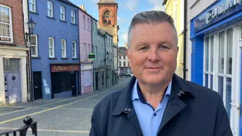 A man wearing a navy jacket with a light blue shirt underneath. He has grey hair is pushed back off his forehead. He is standing on a street with lots of colourful buildings behind him.