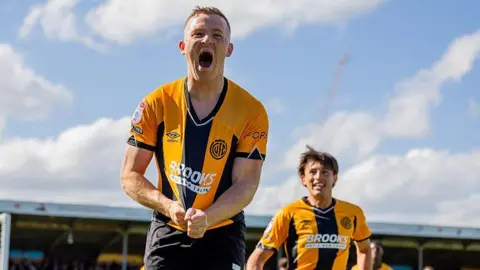Getty Images Shayne Lavery of Cambridge United celebrates scoring a goal. He is running towards the camera with his mouth wide open and clutching his golad and black striped shirt.
