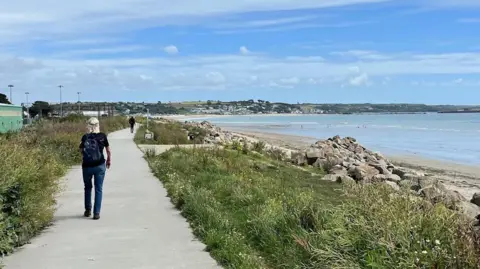 Gail Ward A woman in jeans and a t-shirt walks along a path by a white sandy beach, walking with her back to the camera and a rucksack on her back. 