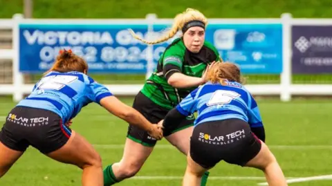 RFU A blonde woman in a green top playing rugby. She had the bball and is about to try passing two opposing players in light and dark blue hooped tops.