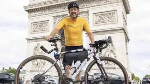 Chris Boardman, who is wearing cycling gear including a helmet, is standing behind his bike and smiling in front of the Arc de Triomphe in Paris. 