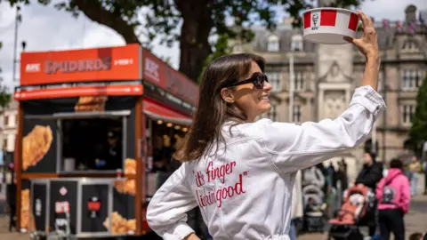 Headland A woman wearing a white jacket with the words 'it's finger-lickin good' holds up a box of KFC