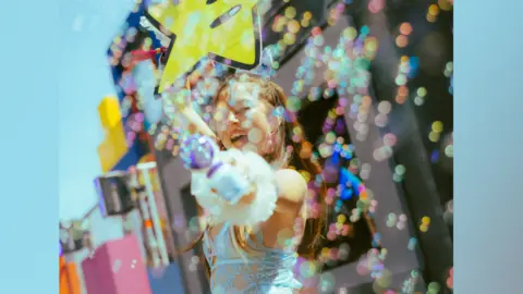 Alastair Brookes A woman holds a yellow smiley star while smiling. She has her eyes closed, with long dark hair down her back and is wearing a blue lace leotard. Iridescent bubbles surround her.