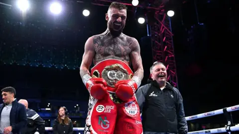 Getty Images Lewis Crocker, a man with dark hair and a beard, tattoos on his chest, red shorts, wearing a red world title belt.