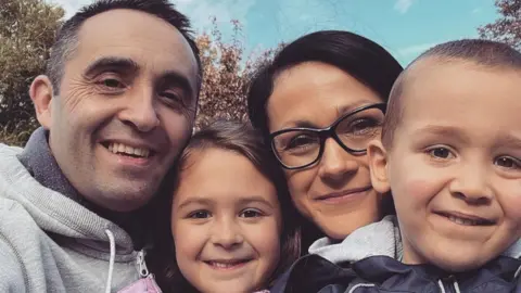 A man with short, dark hair and wearing a grey hoodie smiles while posing for a close-up selfie with his wife, with long dark hair and glasses. His daughter, with brown hair, and his son, with short hair and a navy blue jacket, are also smiling in the image.