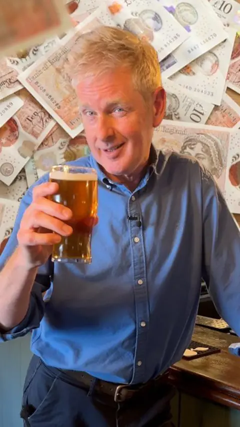 A man in a blue shirt and dark trousers holds a pint in front of a graphic of pound notes
